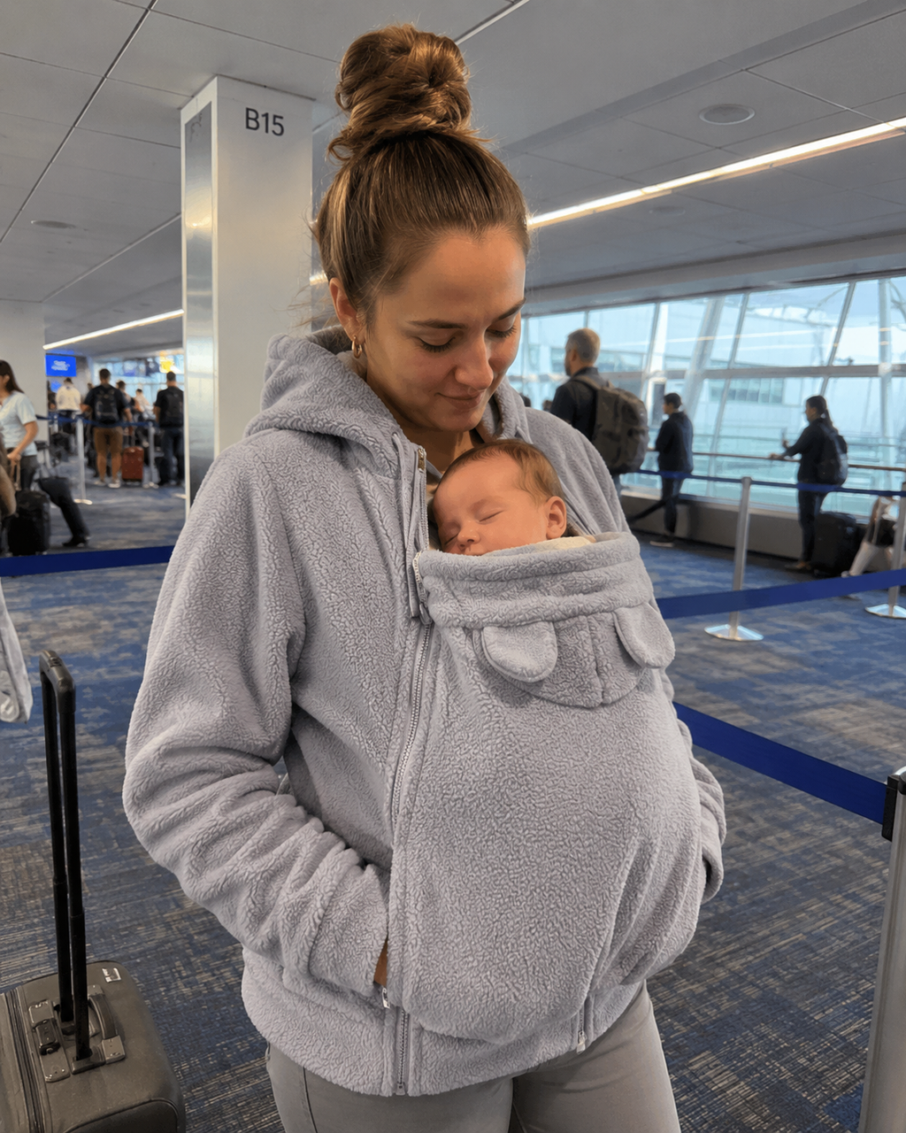 Traveling mom at airport with baby asleep in grey Snoozeroo pouch hoodie