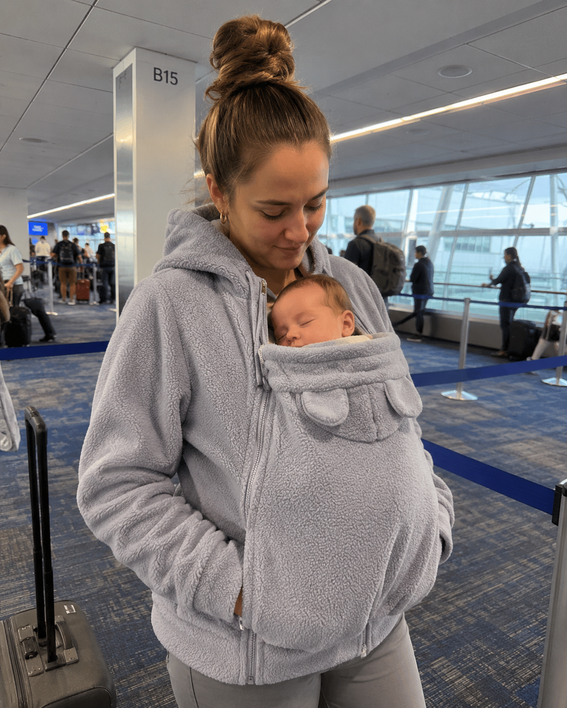 Traveling mom at airport with baby asleep in grey Snoozeroo pouch hoodie